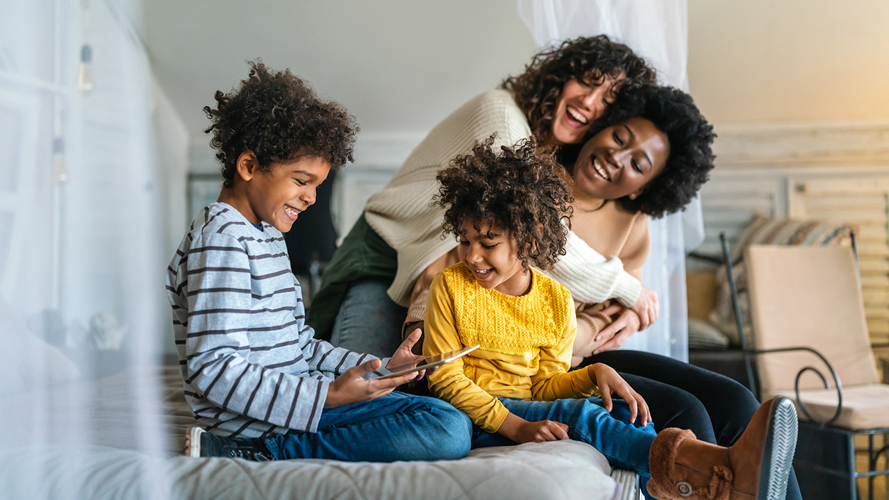 Two moms with their two children smiling and looking at a tablet