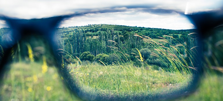 Glasses being held away so a country scene is viewed both clearly through one lens and blurry without it