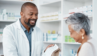 A man in a white coat converses with a woman inside a pharmacy, surrounded by shelves of medication and health products.