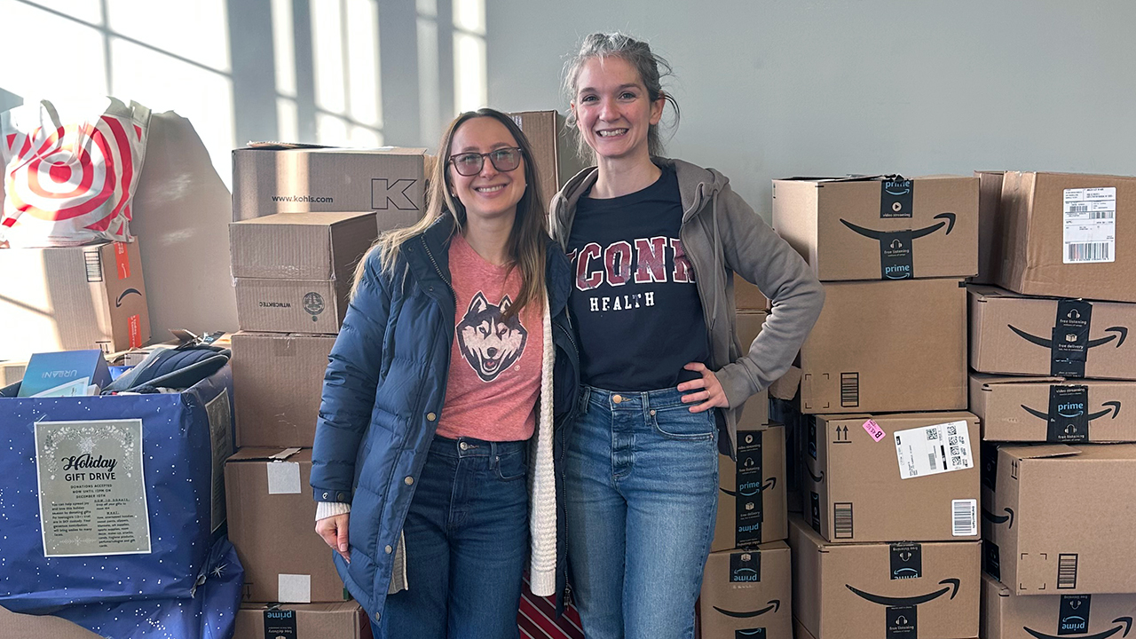 Two ambulatory social workers, Rachel Boxwell and Eleanor Szmurlo, standing in front of Amazon boxes for a holiday toy drive.